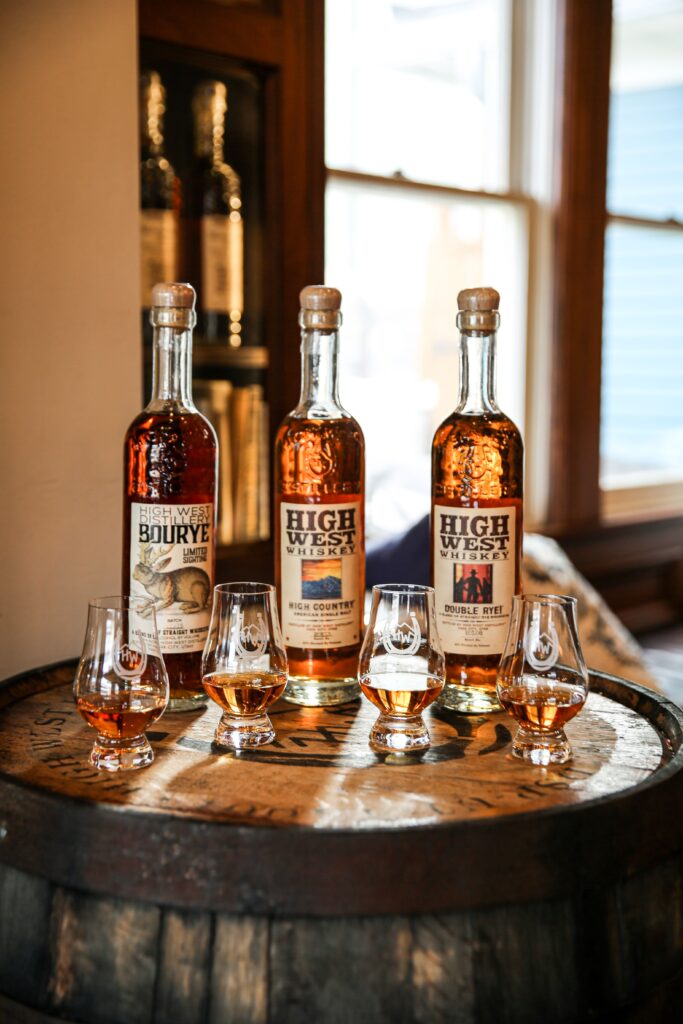 Three bottles of High West Whiskey—Bourye, High Country American Single Malt, and Double Rye—arranged on a rustic wooden barrel alongside four Glencairn tasting glasses filled with amber whiskey.