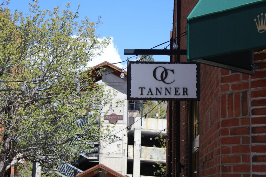 A white rectangular hanging sign for O.C. Tanner jewelry store on a brick building, framed by green trees and a clear blue sky in Park City.