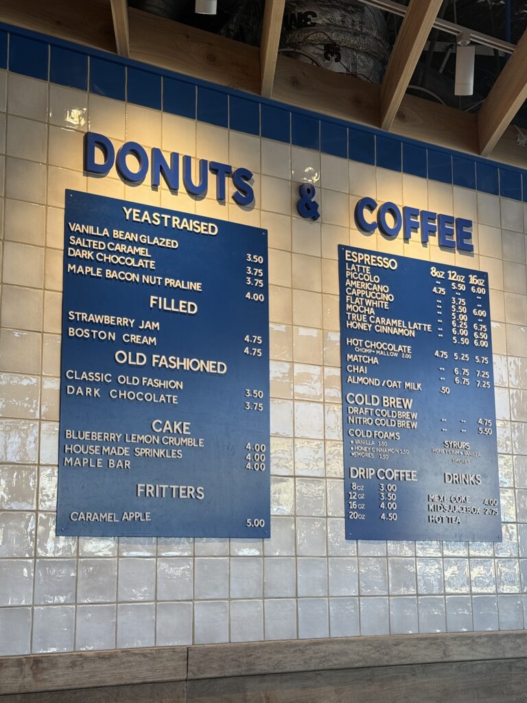A large, blue menu board mounted on a white tiled wall inside a shop. The menu lists various doughnut categories like "Yeast Raised," "Filled," and "Cake," alongside an extensive coffee and espresso menu including lattes, cold brews, and specialty "cloud" drinks.