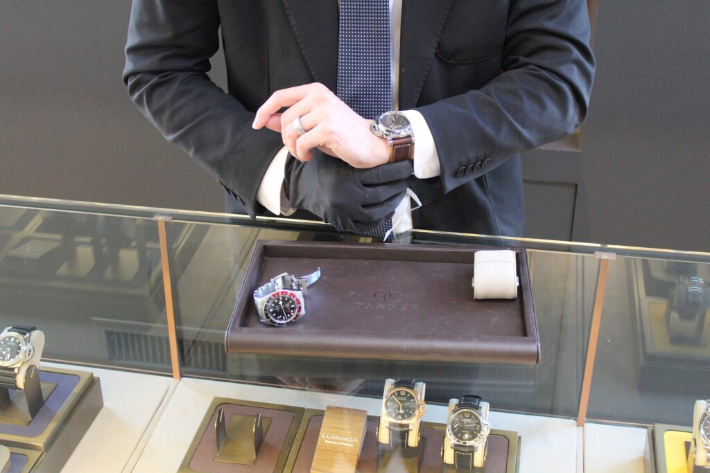 A close-up of a jeweler in a black suit and gloves displaying luxury watches on a leather tray inside an O.C. Tanner showroom.