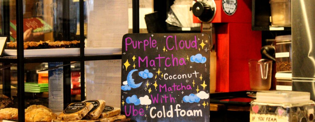 A decorative chalkboard sign reads "Purple Cloud Matcha: Coconut Matcha with Ube Cold Foam," adorned with illustrations of stars, clouds, and a crescent moon.