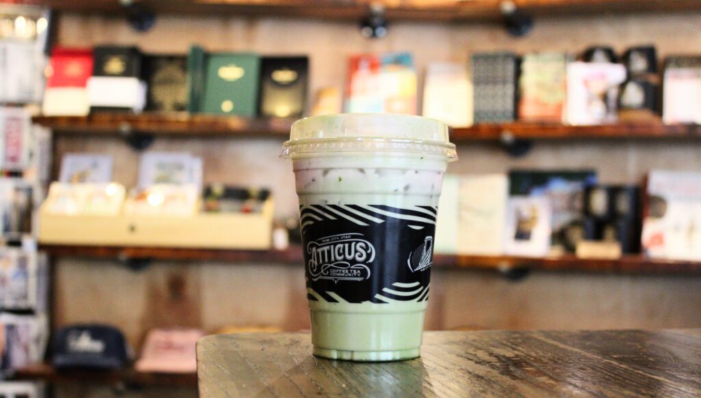 An iced green matcha latte in a clear plastic cup with a black "Atticus Coffee & Tea" sleeve, sitting on a rustic wooden table.