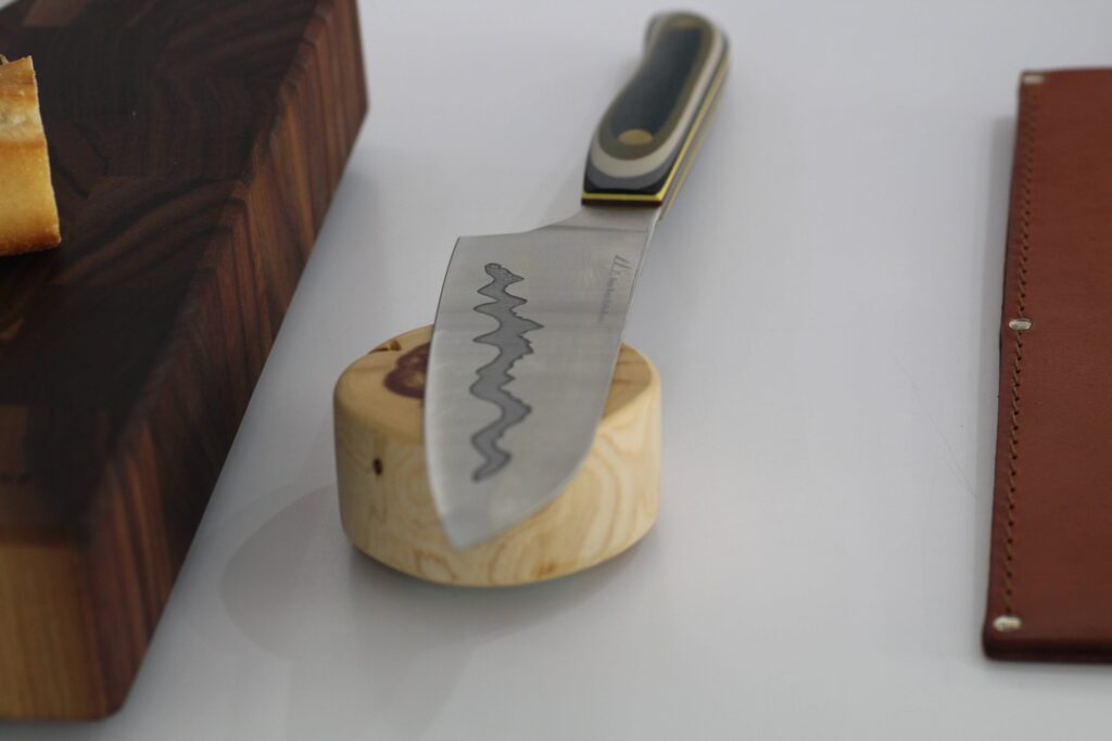 A close-up photograph of a professional-grade kitchen knife with a distinctive wavy-patterned steel blade and a striped multi-color handle, balancing on a light wooden puck. It sits on a white surface with a dark wood cutting board and bread in the background.