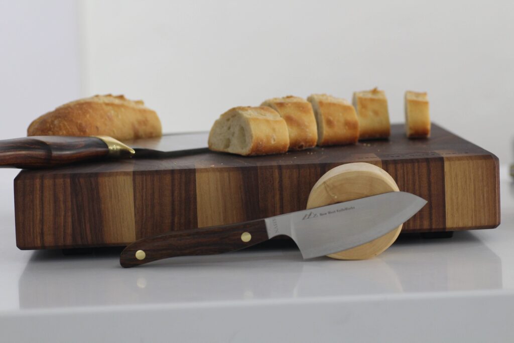 A small utility knife with a dark wooden handle, engraved with "NEW WEST KNIFEWORKS," is positioned in front of a substantial, dark-grained butcher block. A partially sliced baguette rests on the block, with a whole baguette to the side.