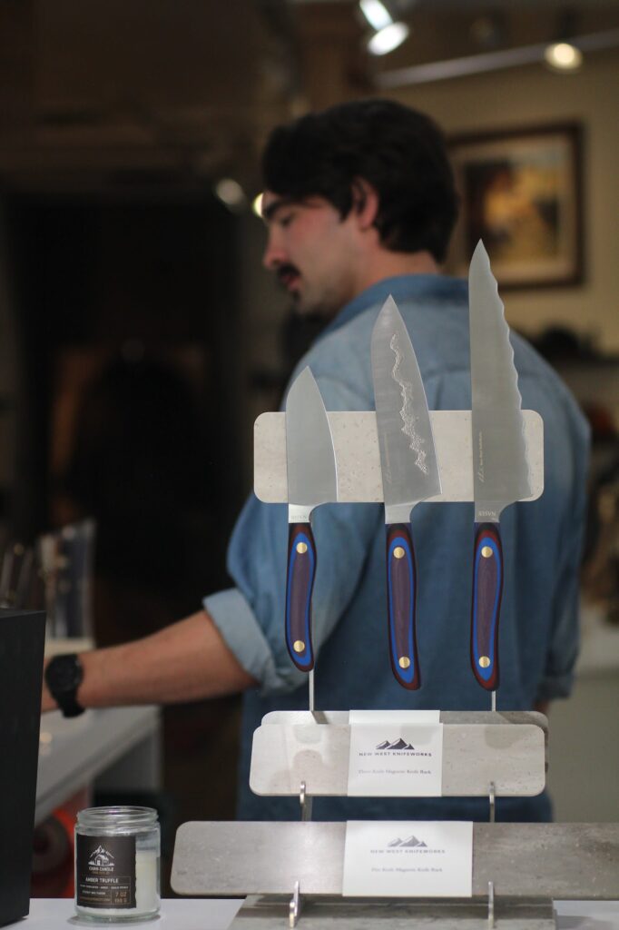 Three kitchen knives with distinctive multi-colored handles (purple, blue, and brown) are displayed vertically on a stone and metal magnetic stand. A man with a beard, wearing a denim shirt, stands blurred in the background, looking at other items in the shop.