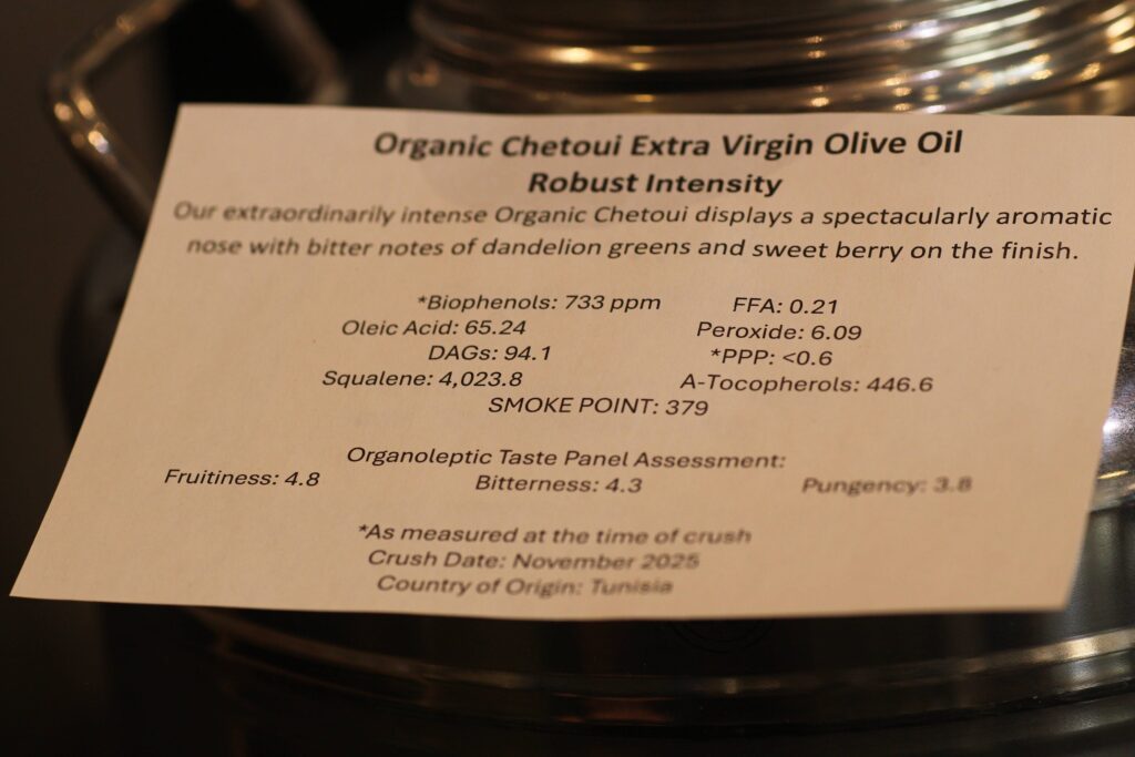 A detailed information card for "Organic Chetoui Extra Virgin Olive Oil" resting on a stainless steel fusti. The card describes the oil as having a "Robust Intensity" with notes of dandelion greens and sweet berry. It lists chemical analysis stats including Biophenols at 733 ppm, a smoke point of 379°F, and a crush date of November 2025 from Tunisia.
