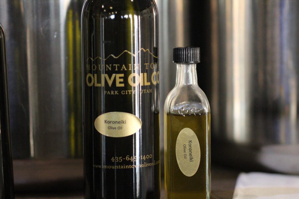 A close-up of two dark glass bottles from the "Mountain Town Olive Oil Co." in Park City, Utah. The larger bottle and a smaller sample-sized bottle both feature gold oval labels identifying the contents as "Koroneiki Olive Oil." The background shows blurred stainless steel containers.