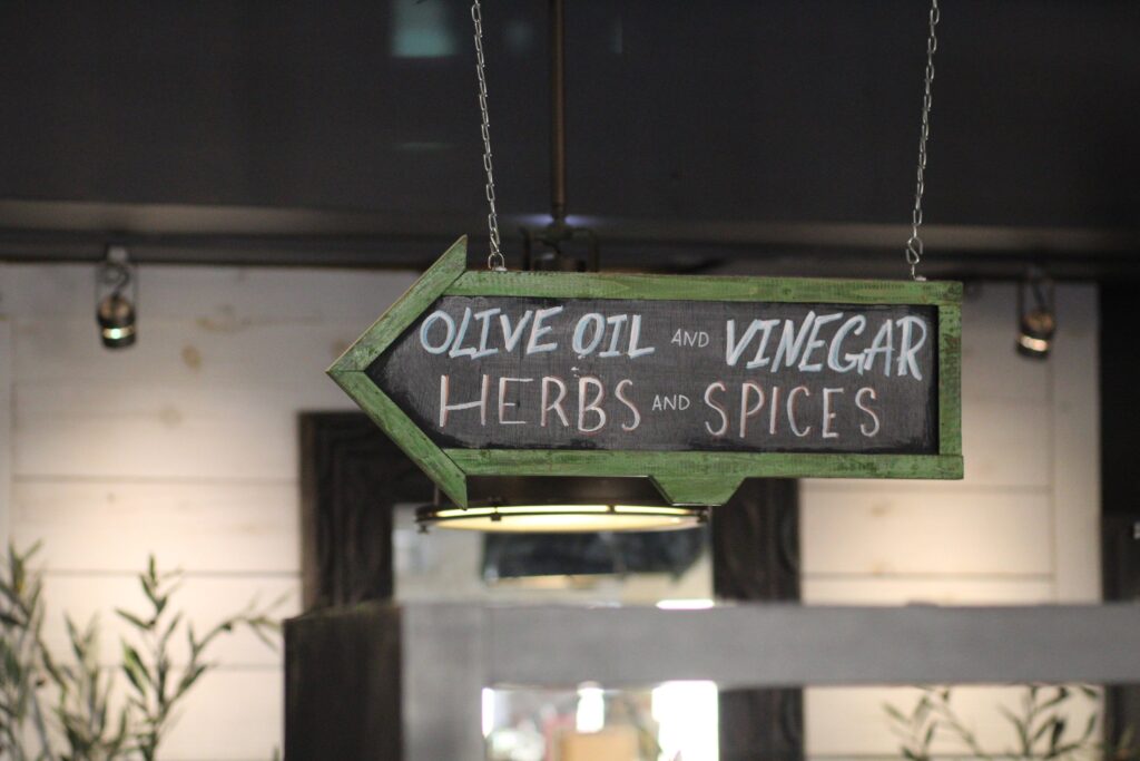 A wooden, arrow-shaped chalkboard sign hanging from thin metal chains in a store. Hand-written in white and light blue chalk, the sign reads "Olive Oil and Vinegar" on the top line and "Herbs and Spices" on the bottom line. The arrow points toward the left.