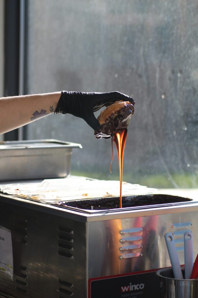 A close-up, action shot of a hand in a black glove dipping a fresh doughnut into a container of thick, glossy chocolate glaze. A long, smooth stream of chocolate drips back into the stainless steel warmer.