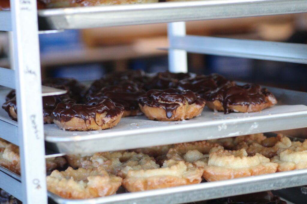A metal bakery rack filled with multiple trays of freshly glazed doughnuts. The middle tray features doughnuts covered in a rich, dark chocolate coating, while the bottom tray holds golden, honey-glazed "old fashioned" style doughnuts.