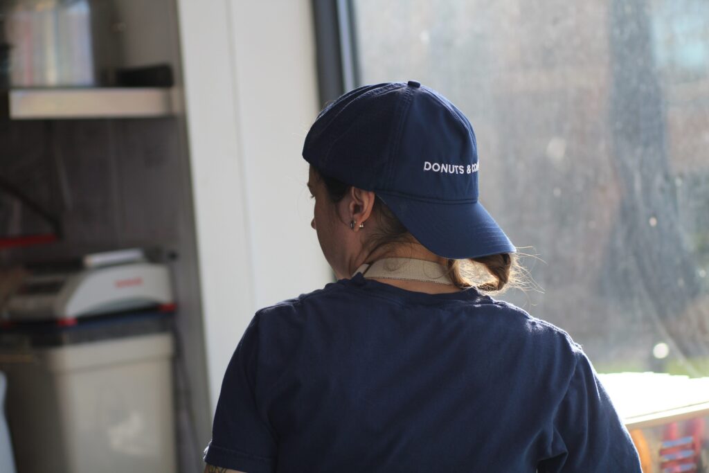 A person viewed from behind wearing a navy blue t-shirt and a matching navy blue baseball cap with "DONUTS & CO" embroidered in white. They are standing in a bright kitchen or shop environment, looking toward a window.