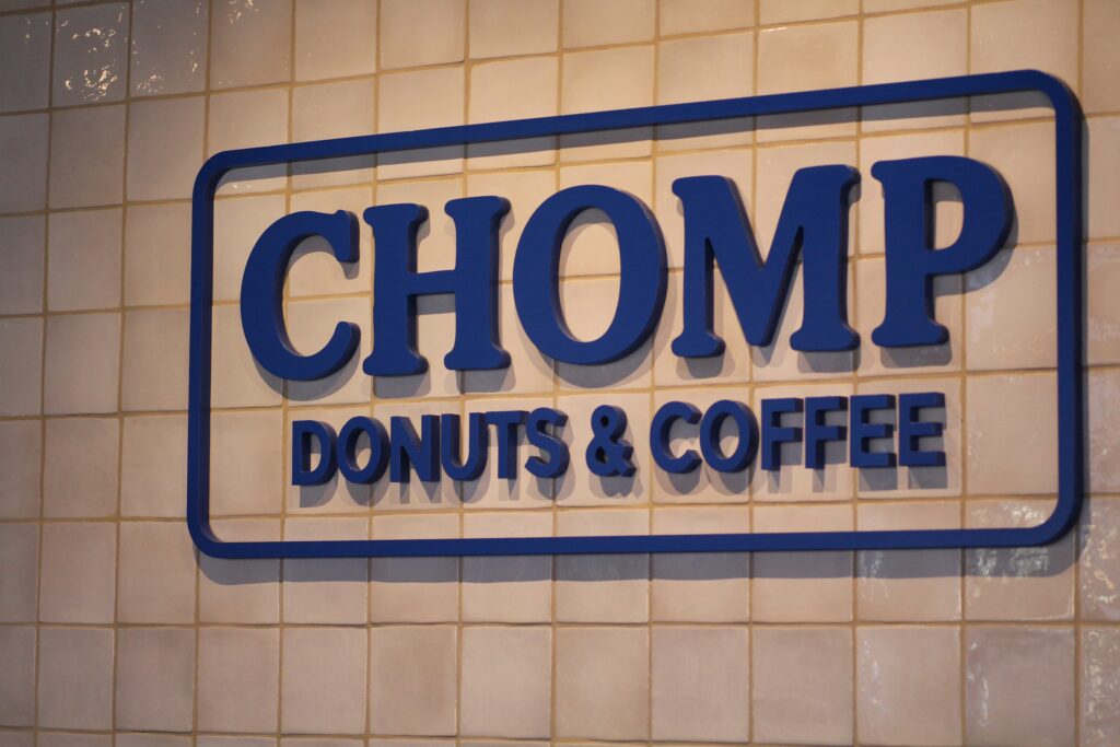 A blue and white "CHOMP DONUTS & COFFEE" sign mounted on a wall of off-white square tiles. The sign features bold, blue 3D lettering enclosed in a rounded blue rectangular border.