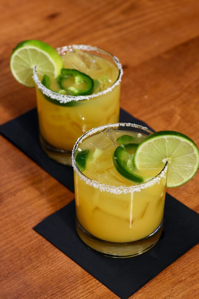 Two bright orange mango margaritas served in glasses with a sea salt The cocktails are garnished with fresh jalapeño slices and lime wedges, showing a vibrant contrast between the tropical fruit color and the spicy green accents.