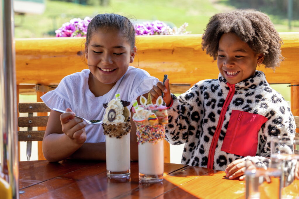 Two young girls smiling and enjoying extravagant, fully-loaded milkshakes on an outdoor mountain deck, highlighting family-friendly options for Deer Valley dining.