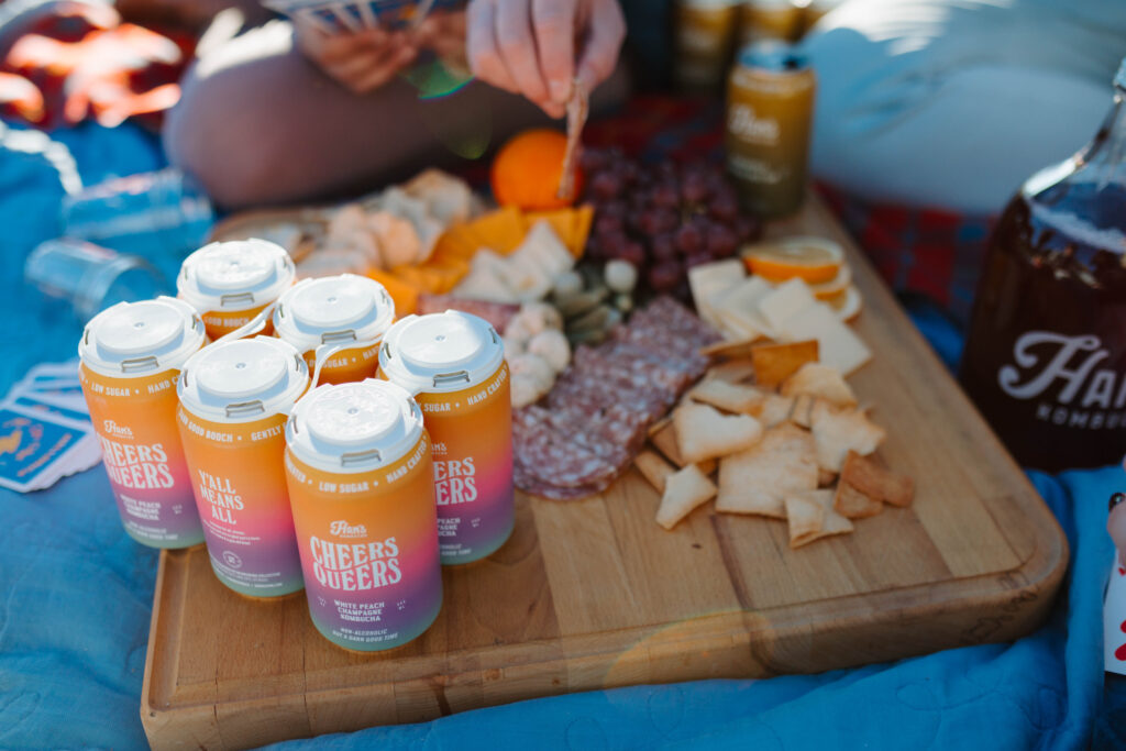 Han's Kombucha cans, including the "Cheers Queers" white peach champagne flavor, served on a picnic blanket alongside a gourmet charcuterie board with cheese, crackers, and meats.