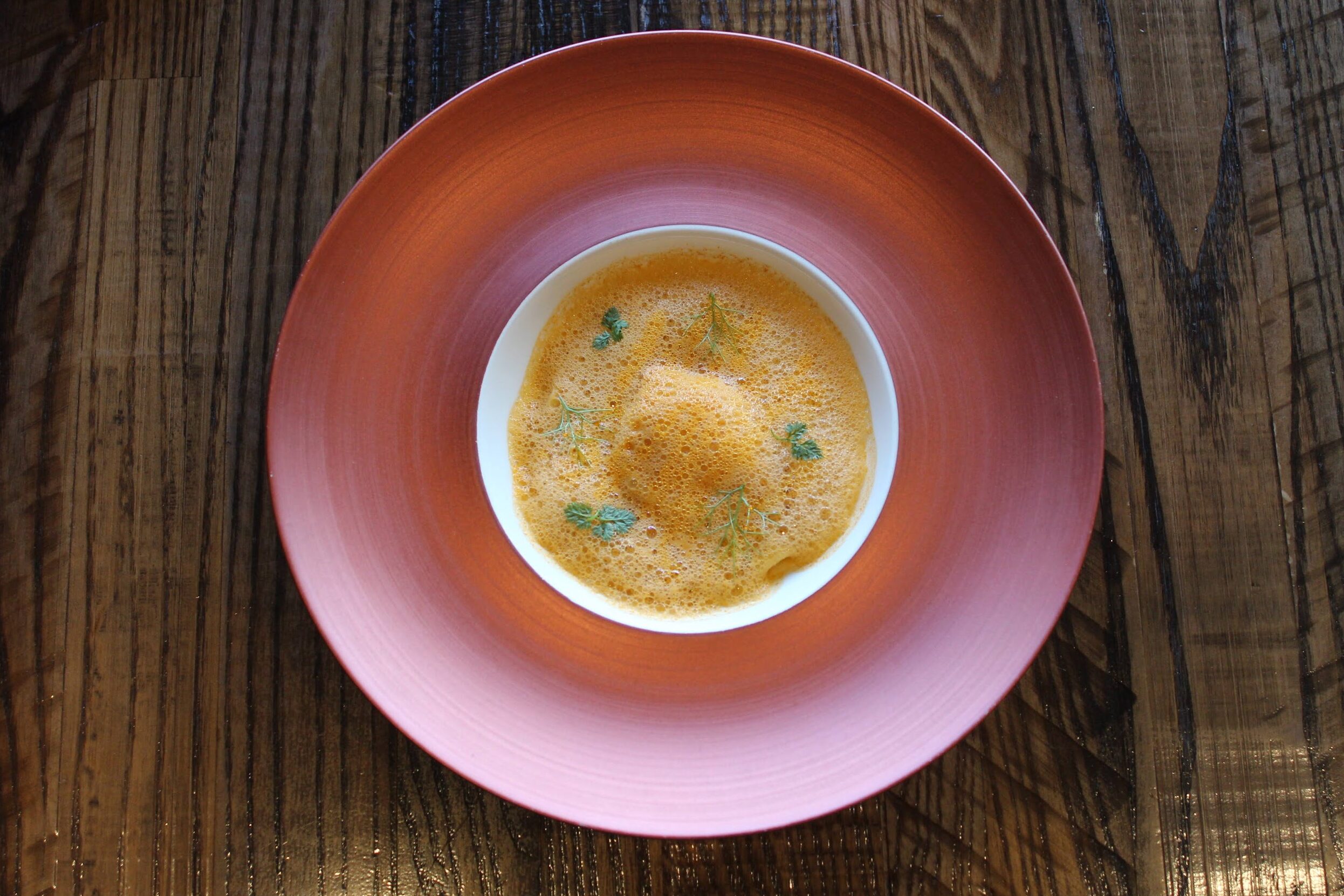 An overhead, high-angle shot of a gourmet soup or foam dish served in a small white bowl, which is centered within a large, wide-rimmed copper-colored plate. The dish is garnished with delicate green herbs and sits on a dark, rustic wooden table with a prominent grain.